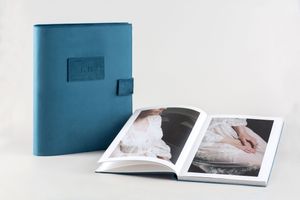 A teal leather photo album lies open displaying wedding dress detail photographs against a white background.