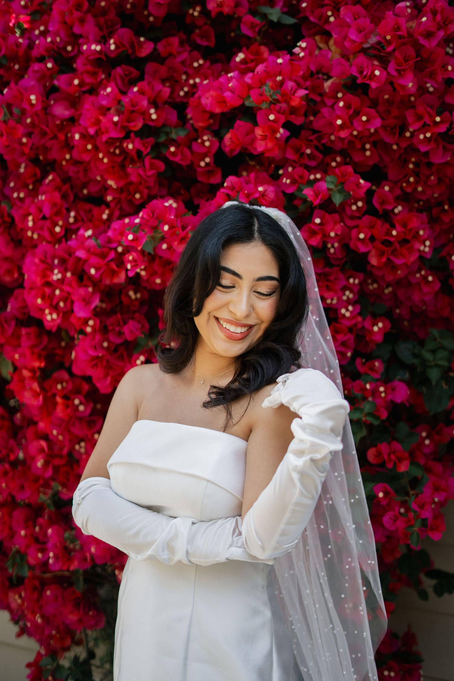 Bride in white wedding dress and gloves poses elegantly against vibrant red bougainvillea flowers in bloom.
