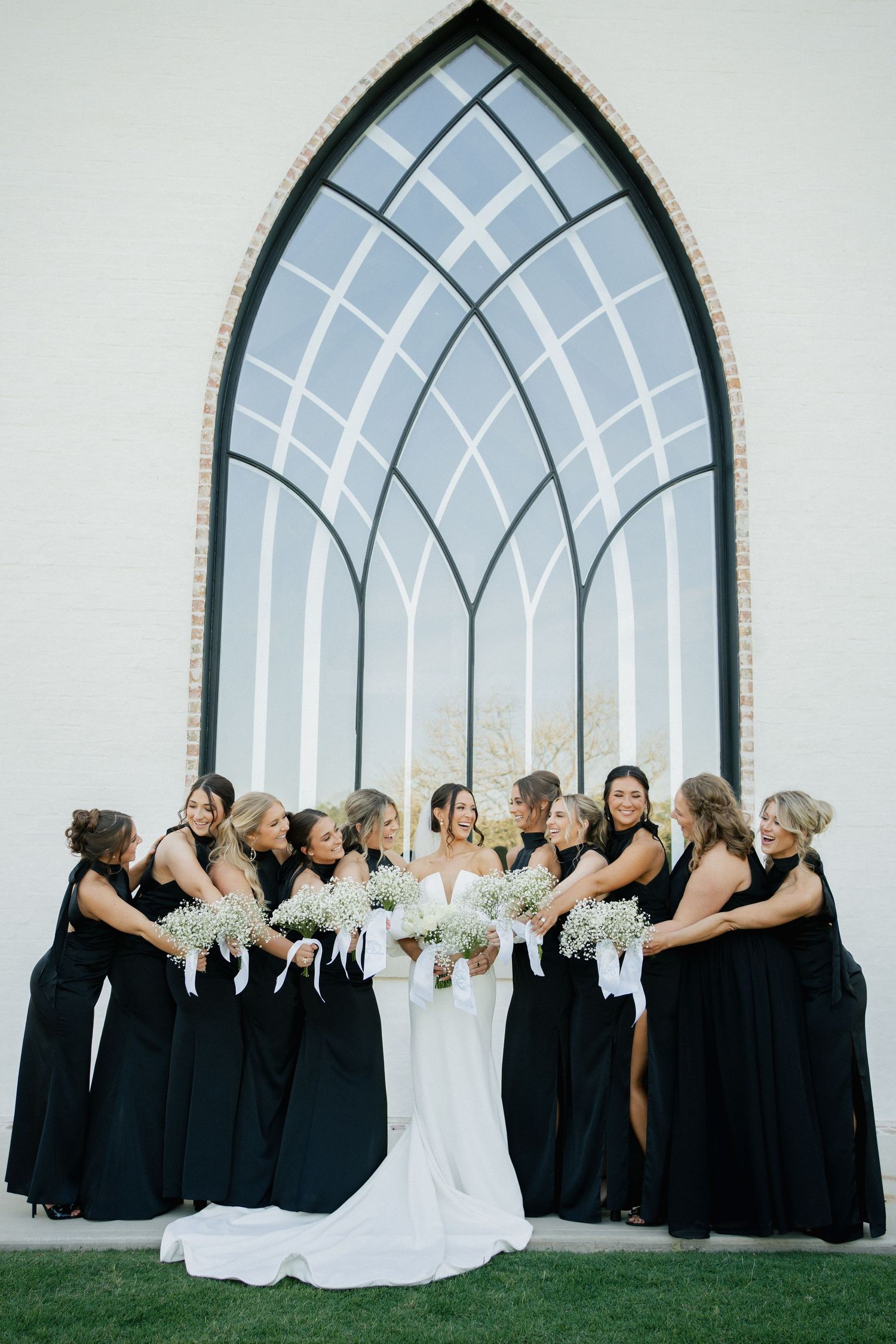 Wedding party gathered in front of a Gothic church window letting natural light illuminate the bride in white and bridesmaids in black.