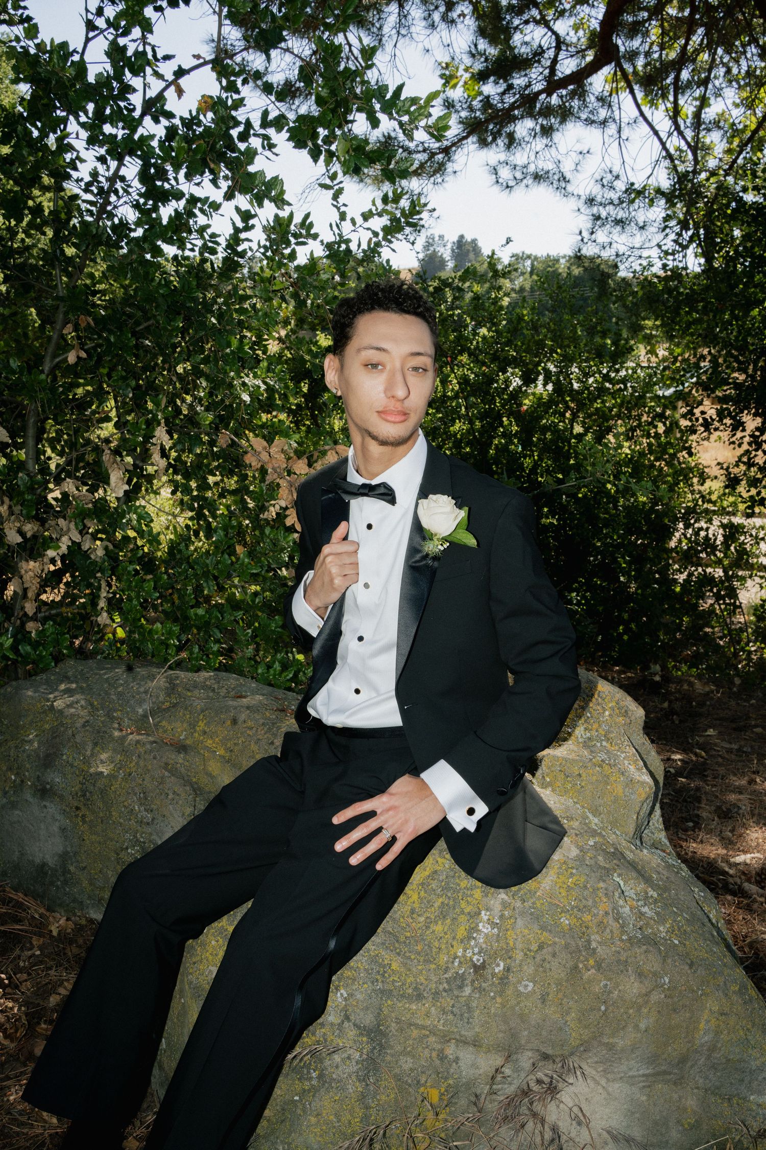 A person in a formal black tuxedo with bow tie and white boutonniere poses on a large rock outdoors among greenery.