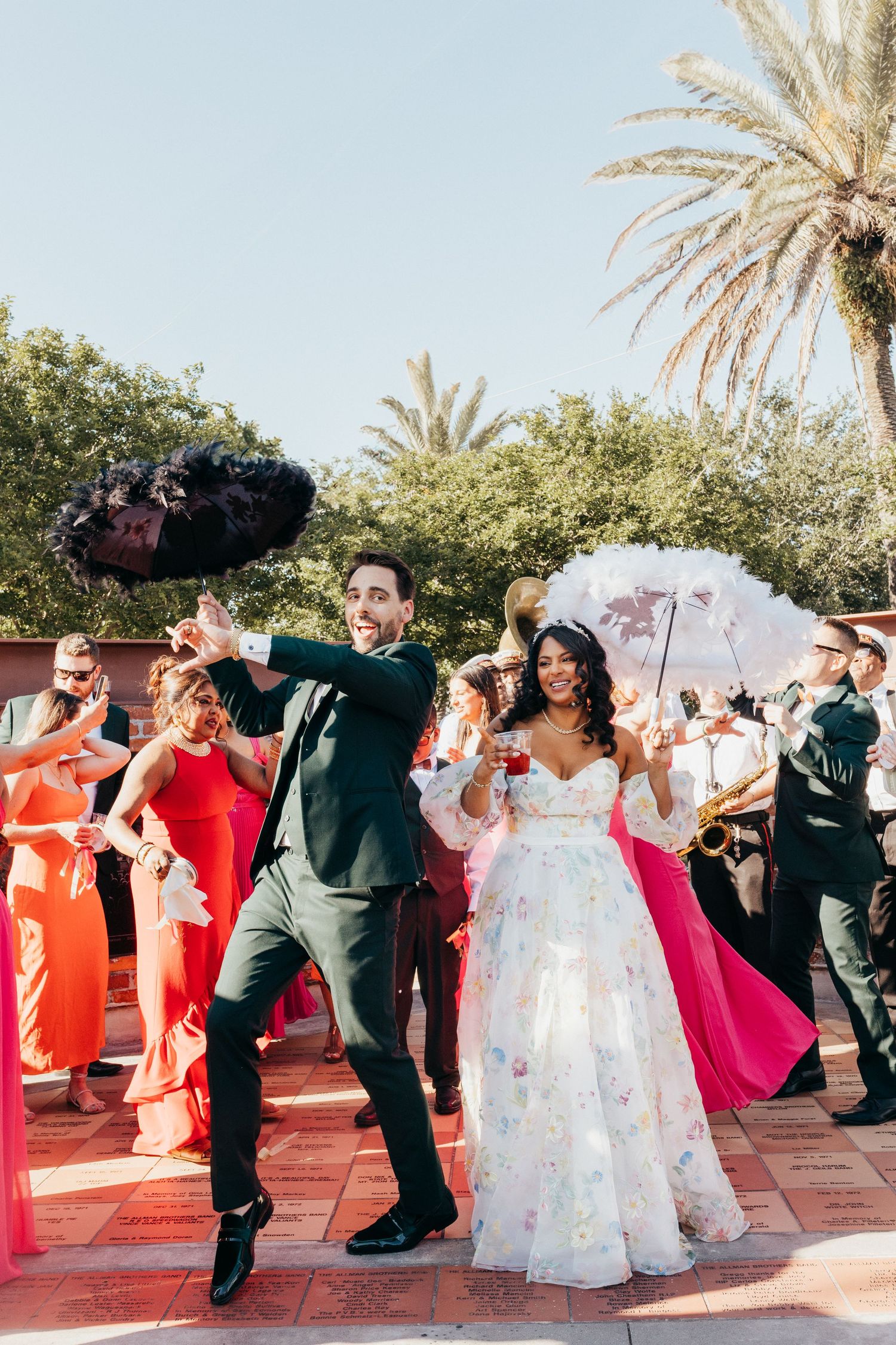 Wedding guests form a joyous conga line on an outdoor dance floor with palm trees in the background.