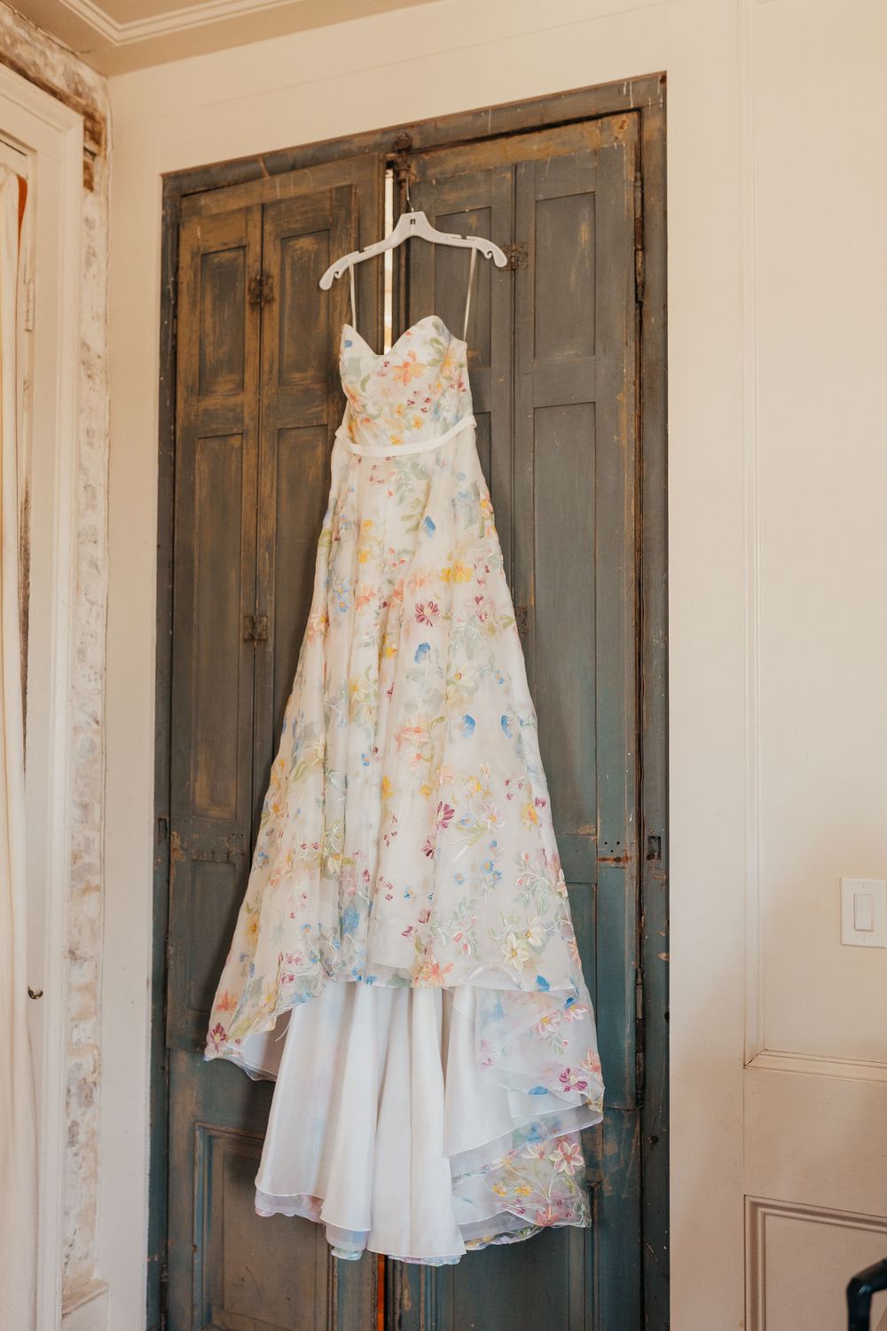 A floral wedding dress with soft pastel colors hangs on a white hanger against a vintage wooden door. house of Savin dress