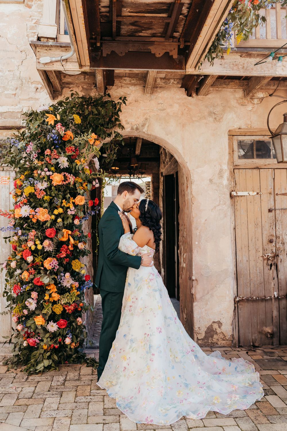 Romantic couple poses in rustic stone villa with vibrant floral arch featuring orange and pink blooms. at race and religious