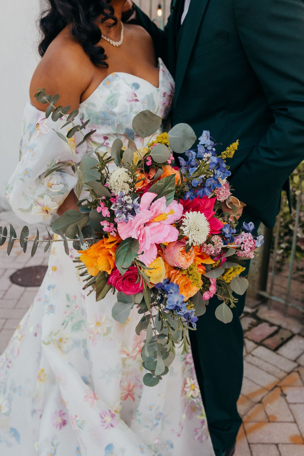 A colorful wildflower wedding bouquet with pink, orange, and purple blooms against a floral print dress and dark suit.