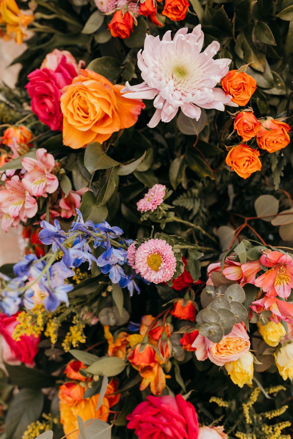 Colorful floral arrangement featuring orange roses, pink chrysanthemums, blue delphiniums and various blooms against dark foliage.