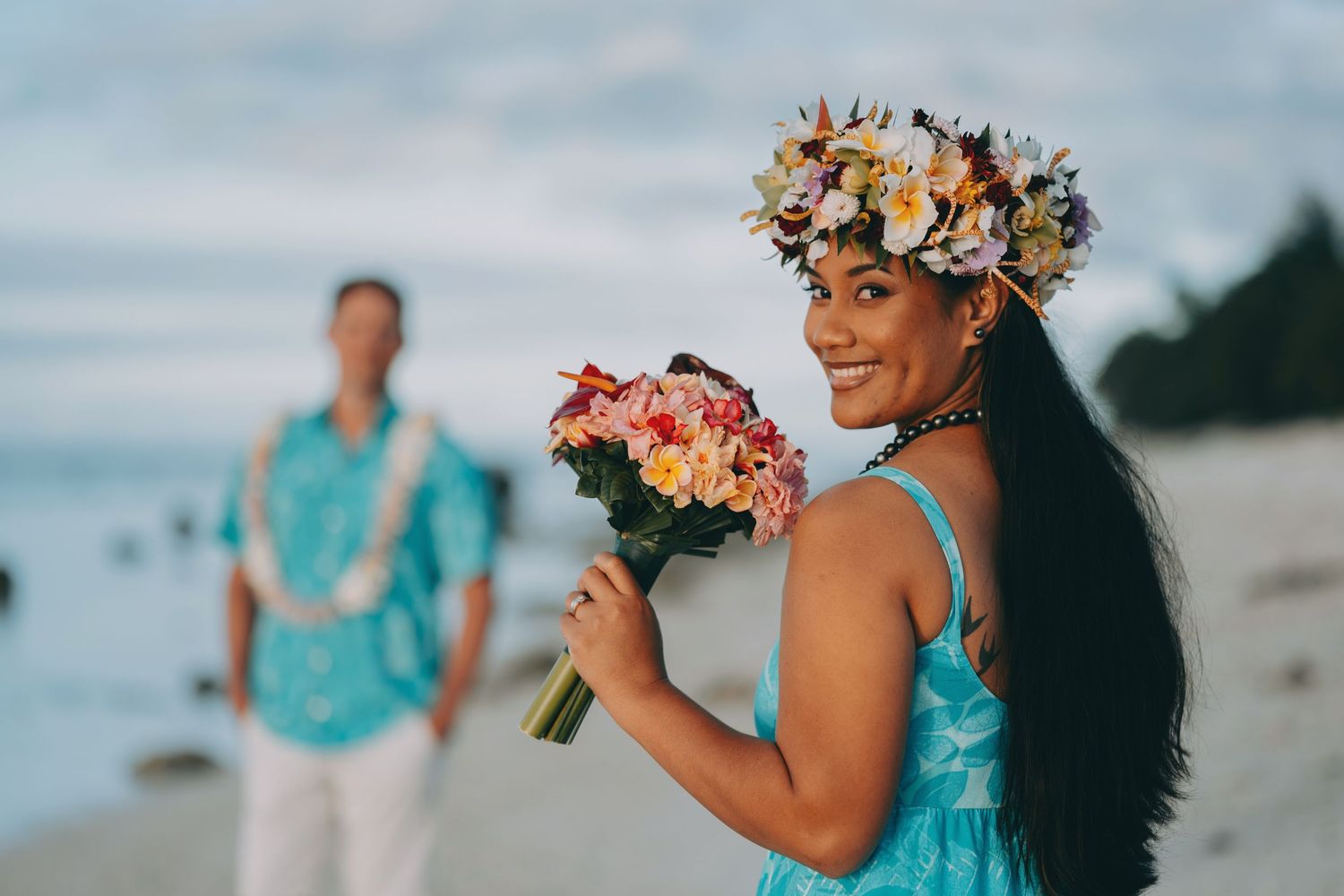 A couple poses for romantic beach engagement photos in coordinating turquoise outfits with tropical flowers.
