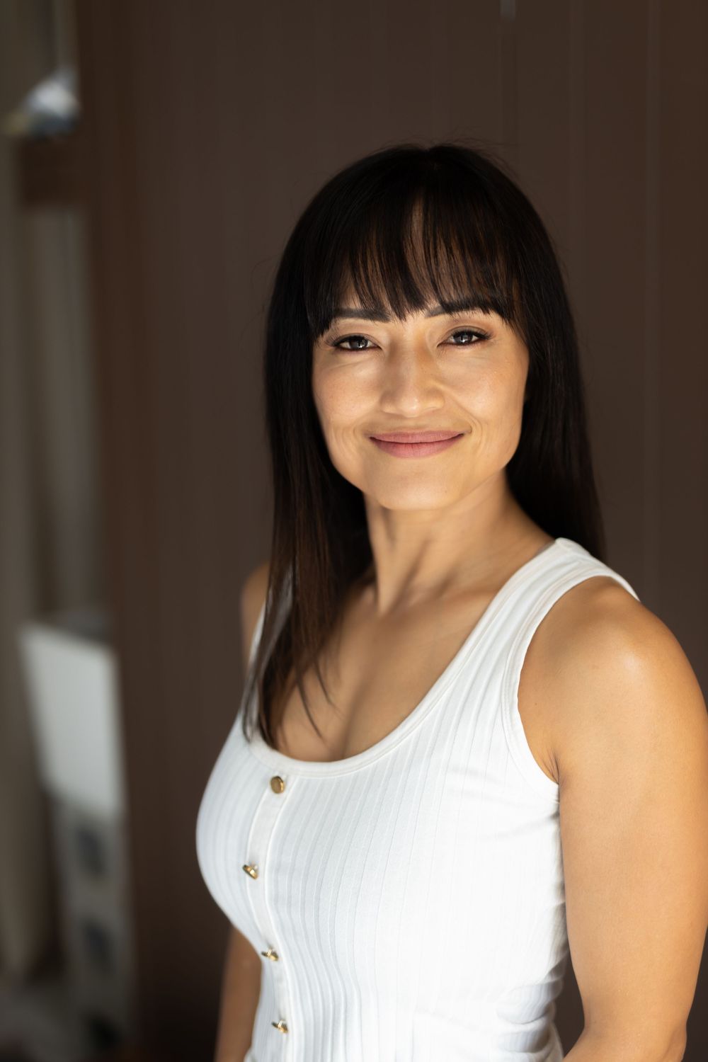 Professional portrait of a woman in a white tank top with buttons smiling warmly against a dark background.