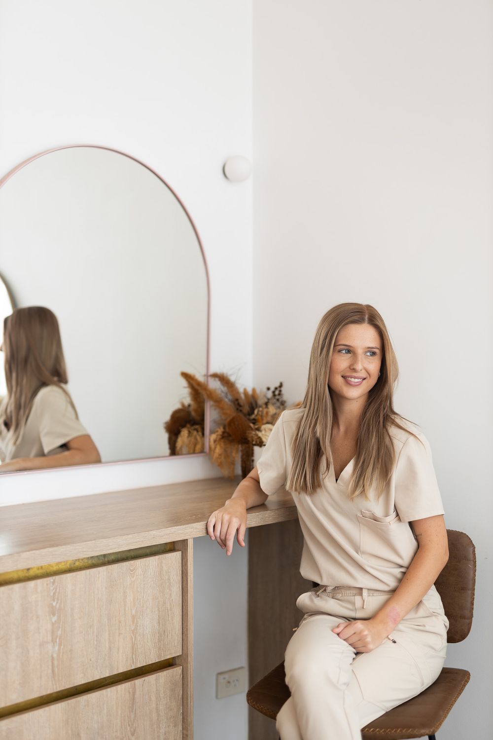 Someone sitting in a brown chair by a vanity mirror and decor in a bright minimal interior space wearing neutral tones.