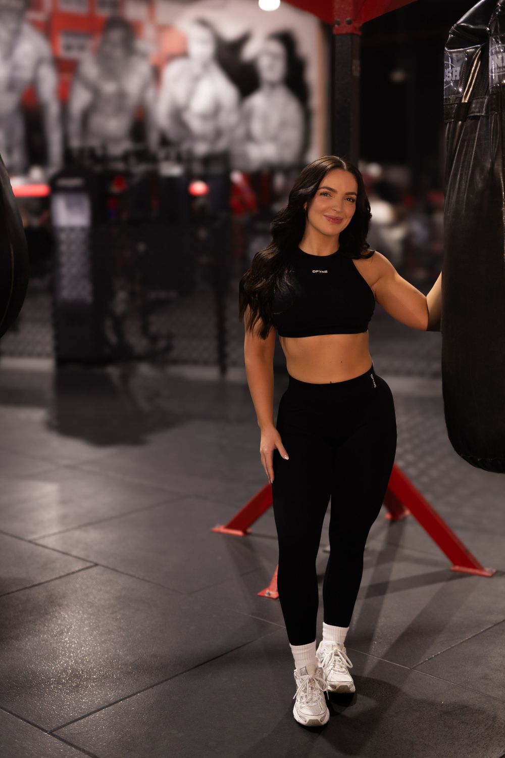 Female athlete in workout gear performs stretching exercises on a dark gym floor with red accents visible.