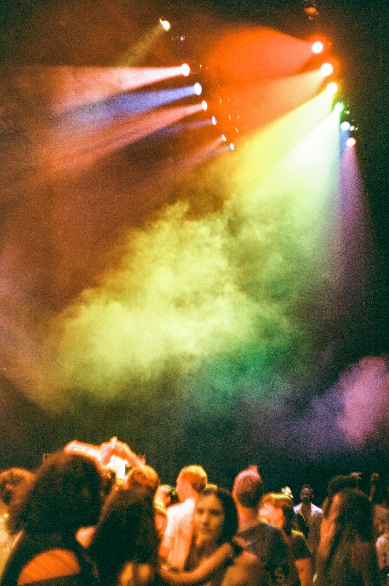 Colorful stage lights illuminate a smoky concert venue with a crowd of silhouetted people dancing below.