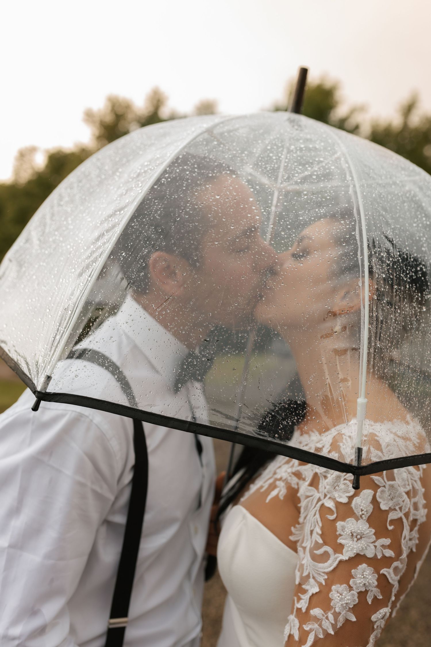 Bride and groom kissing under clear umbrella in rain on wedding day.