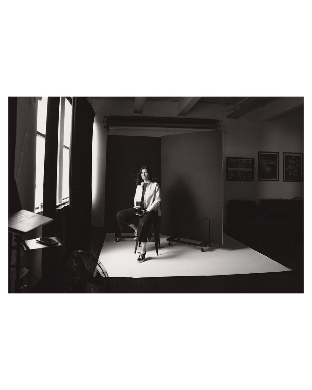 Black and white photo of a figure seated on a bar stool in a studio setting with dramatic window lighting.