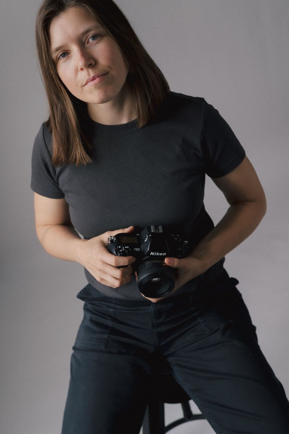 Photographer wearing black t-shirt and pants holds professional camera against gray studio background.