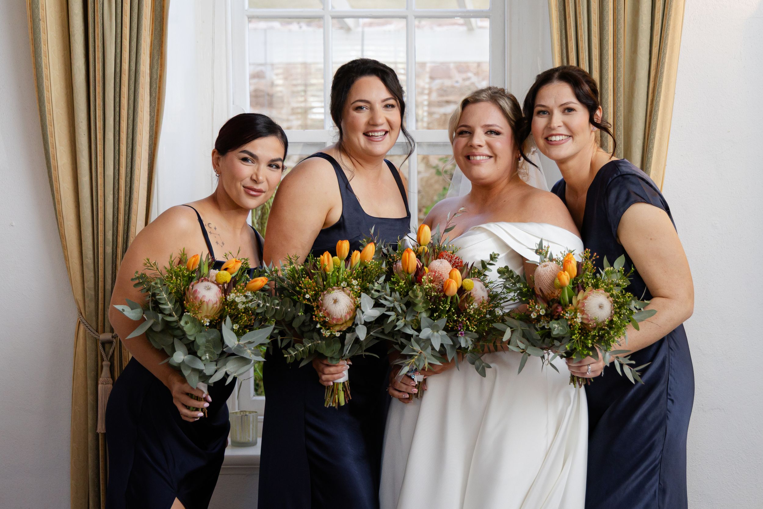 A wedding party with bridesmaids wearing navy blue dresses pose together holding bouquets of flowers in a room.