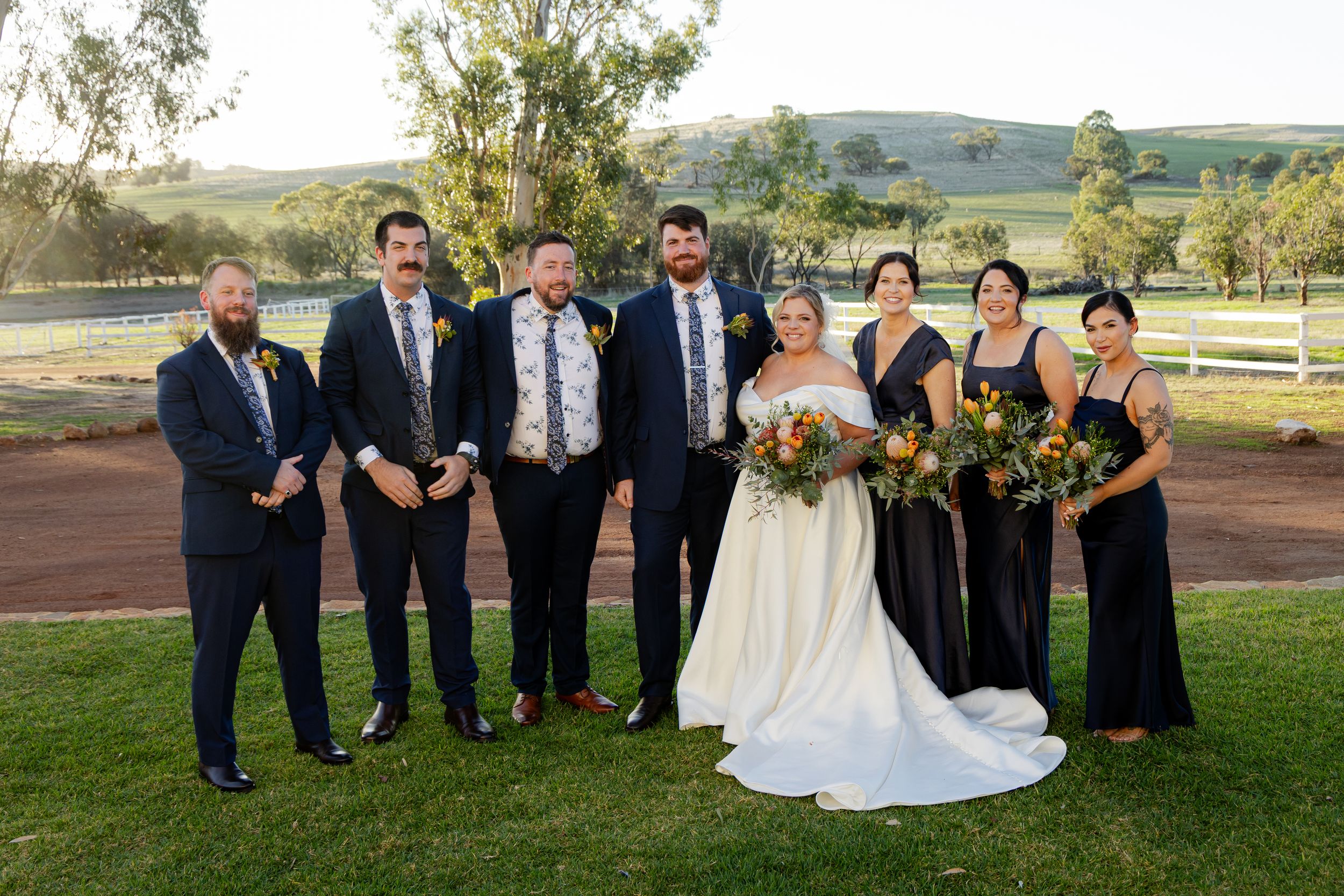 Wedding party members in navy and white attire pose together on a grassy lawn at an outdoor countryside venue.