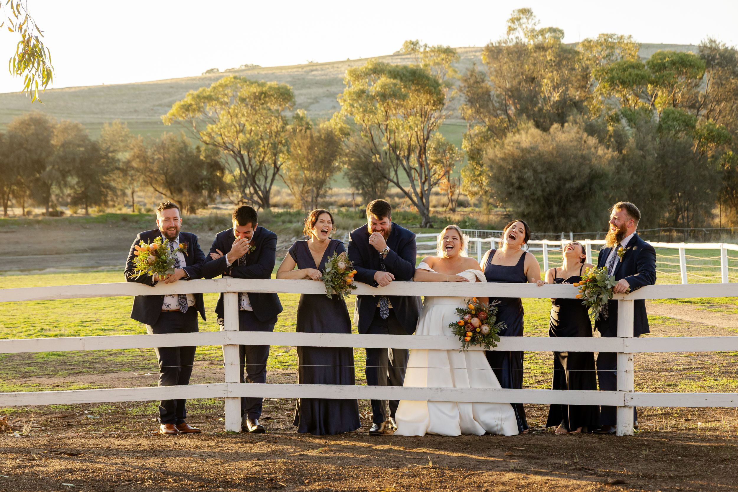 A wedding party poses on a rustic fence at sunset in a rural countryside setting against rolling hills.