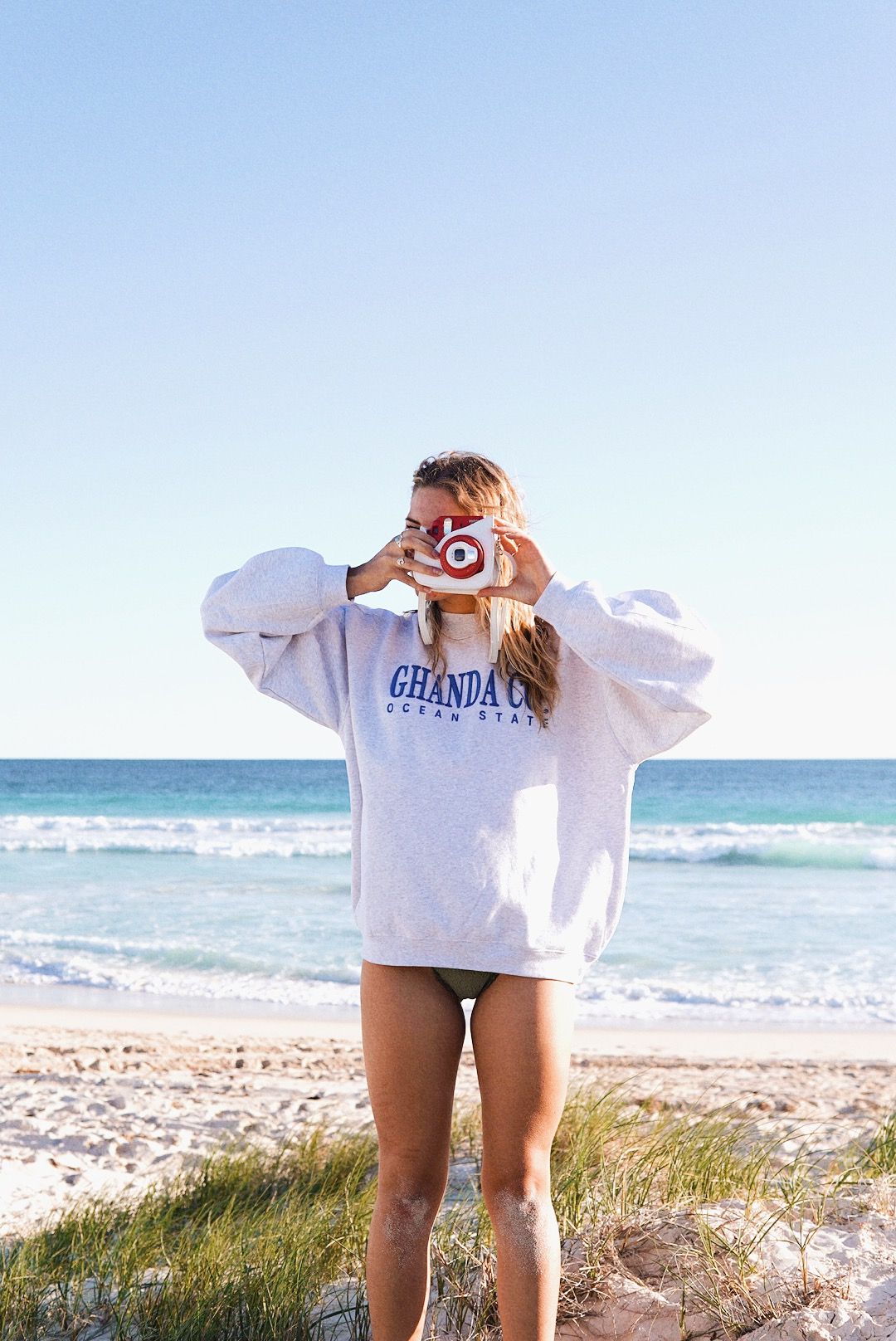 Person in oversized sweatshirt taking photo at beach with waves in background.