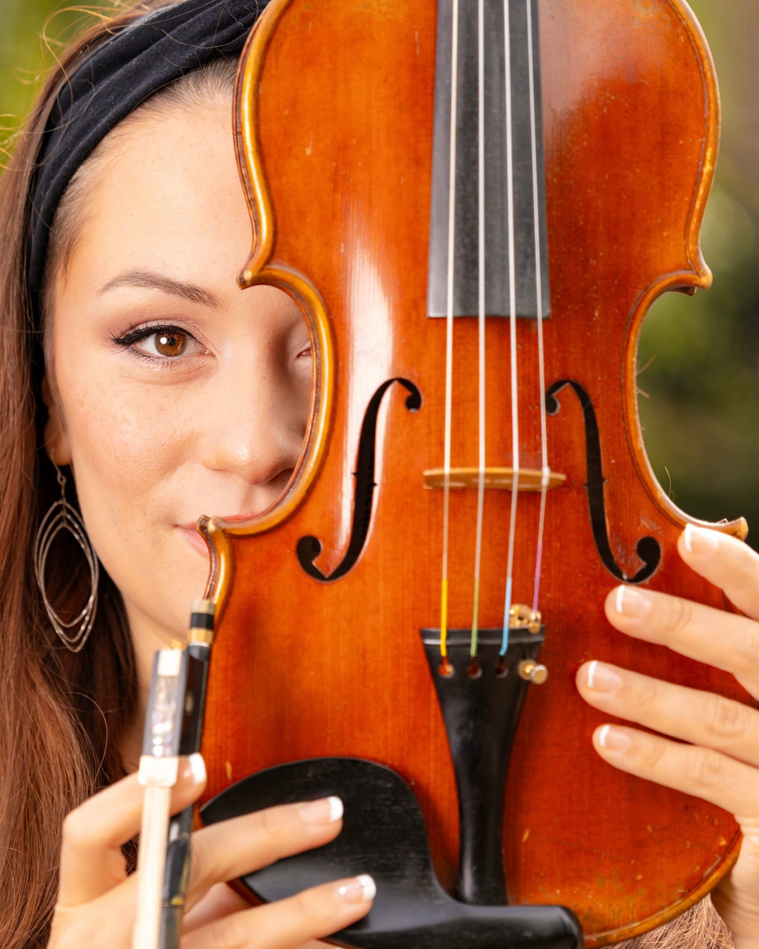 Woman with a dark headband and drop earrings peeks from behind a polished wooden violin while holding a bow in her hand.