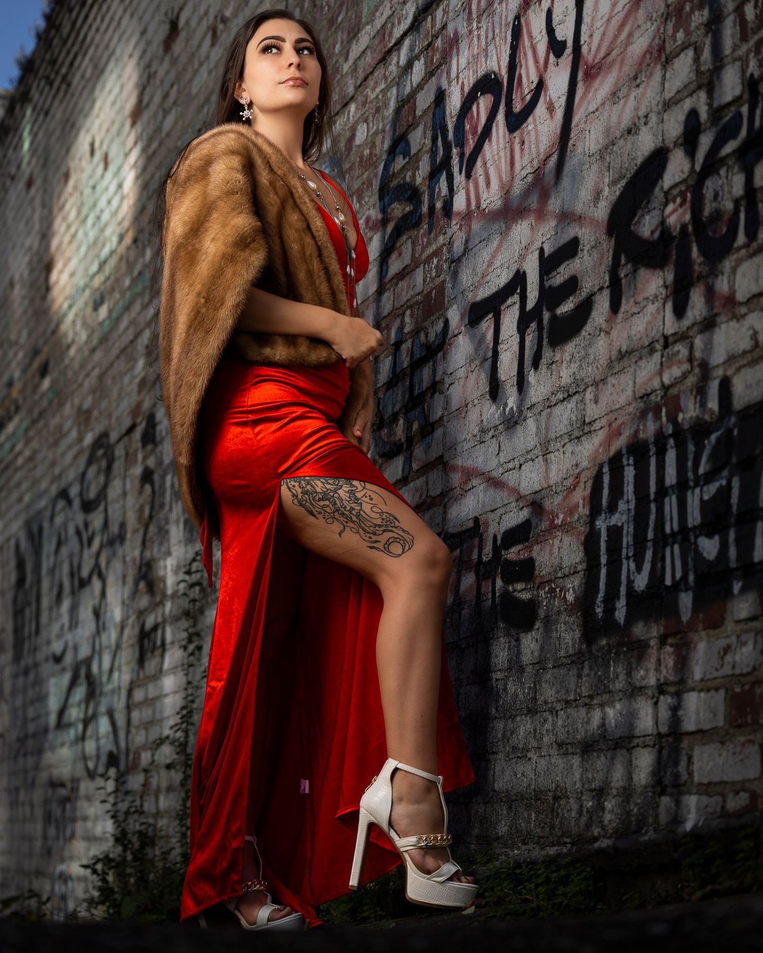 Woman in red satin gown with fur wrap poses against graffiti wall wearing white heels.