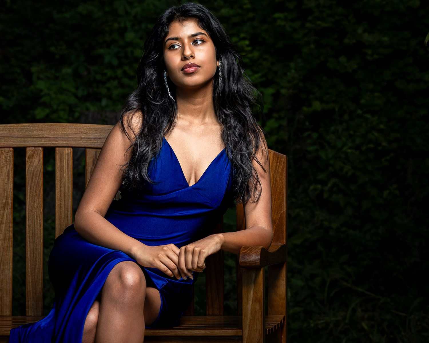 Young woman in elegant blue dress seated on wooden bench outdoors against lush green foliage backdrop.
