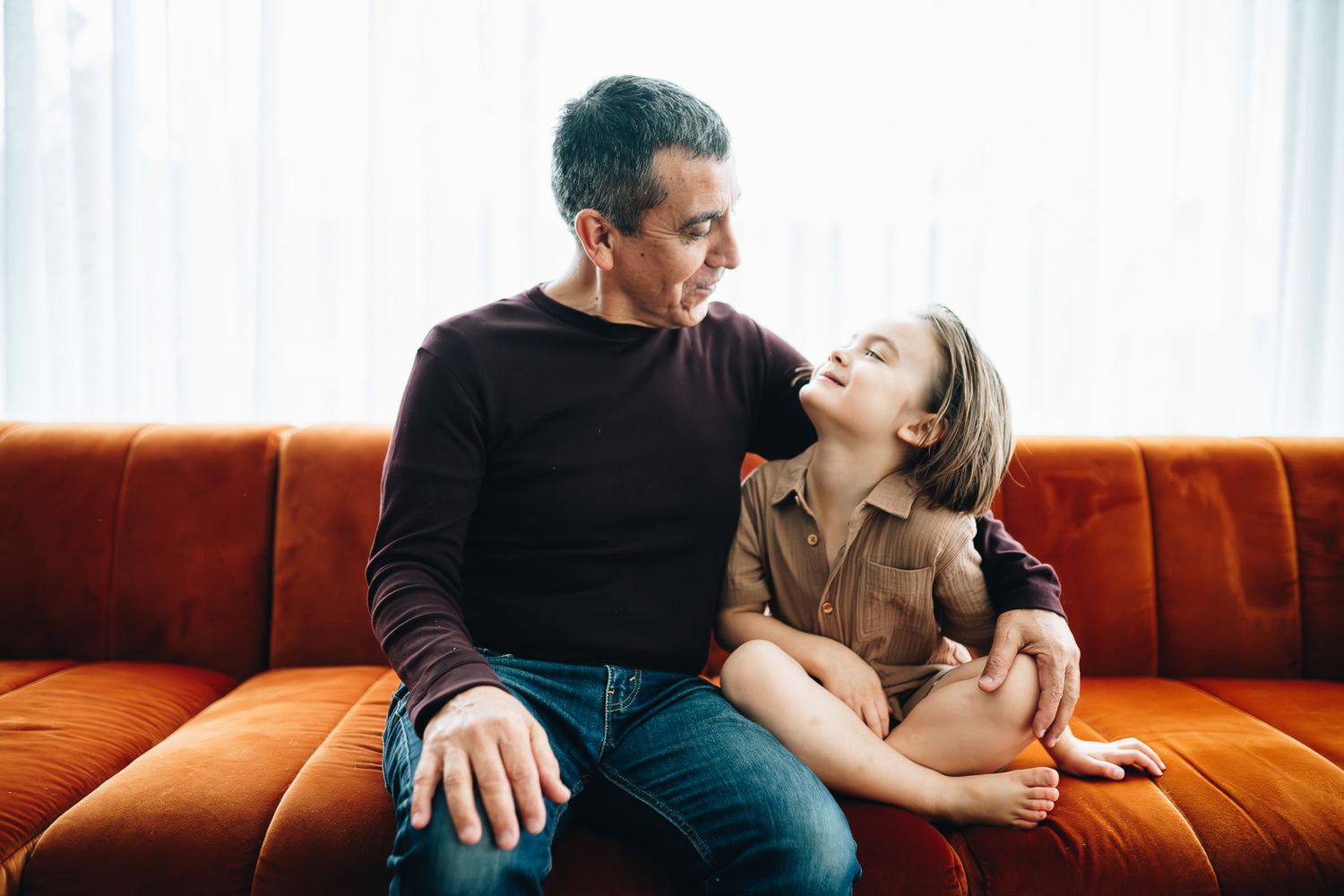 Grandfather and child share a joyful moment of laughter while sitting together on an orange couch taken by Melbourne family photographer