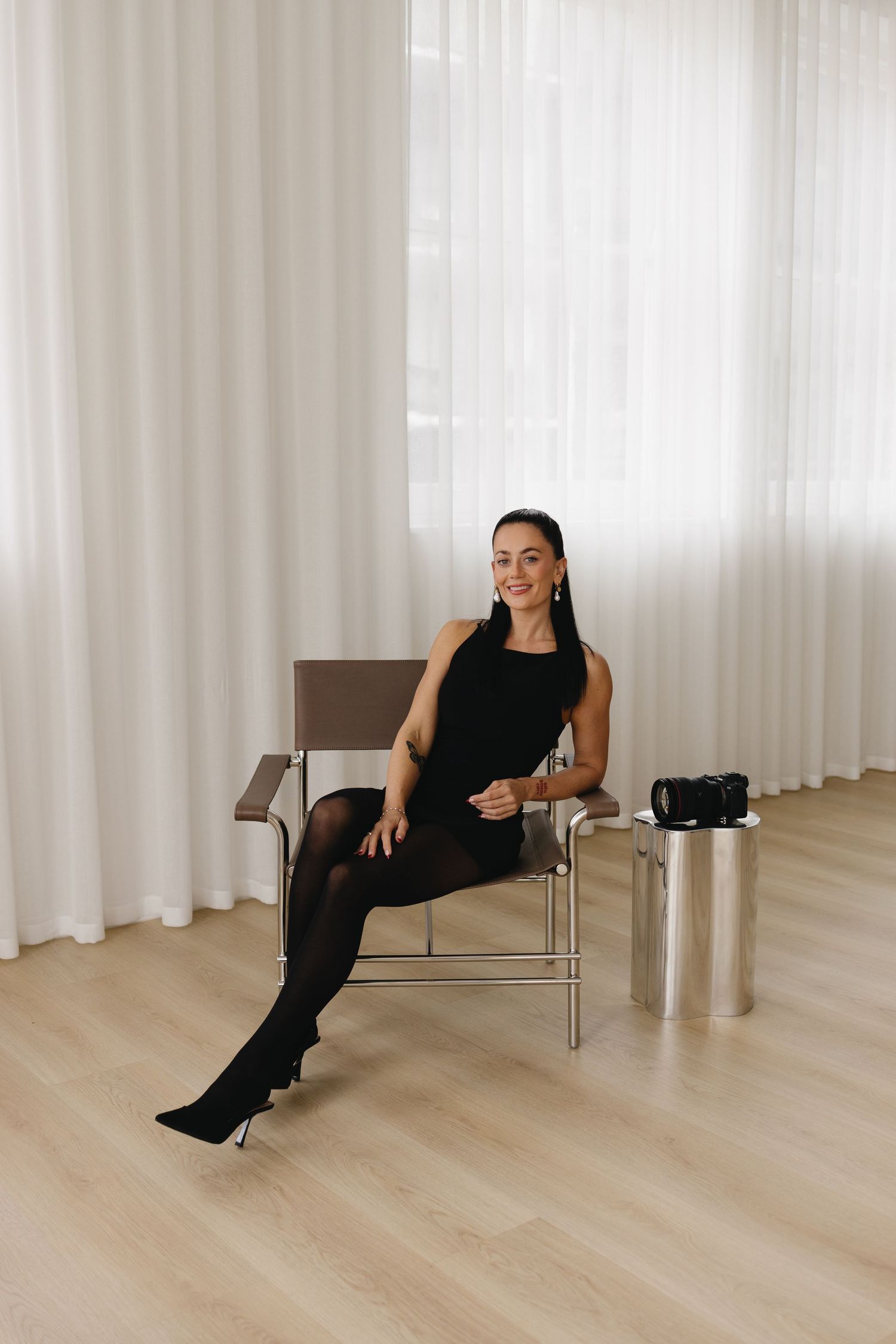 A person in a black strapless top and white pants sits relaxed in a mid-century modern armchair against a white background.