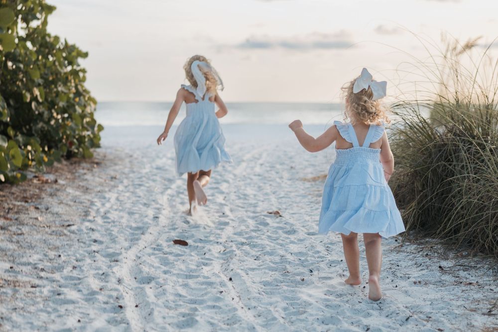 Two figures in matching light blue dresses dance playfully on a sandy beach path during golden hour.