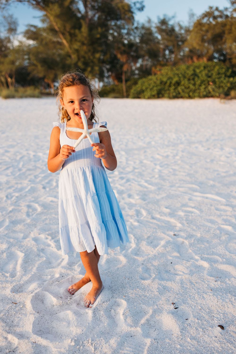 A young child in a white dress holds a starfish while standing barefoot on pristine white sand at the beach.