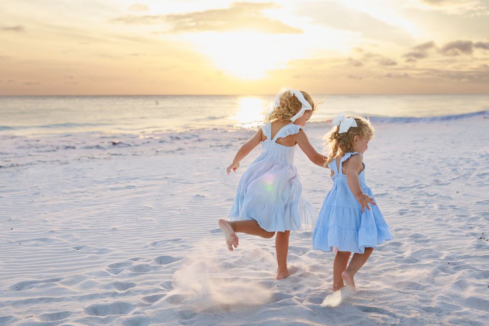 Two people in light blue dresses walk along a pristine white sandy beach at sunset.
