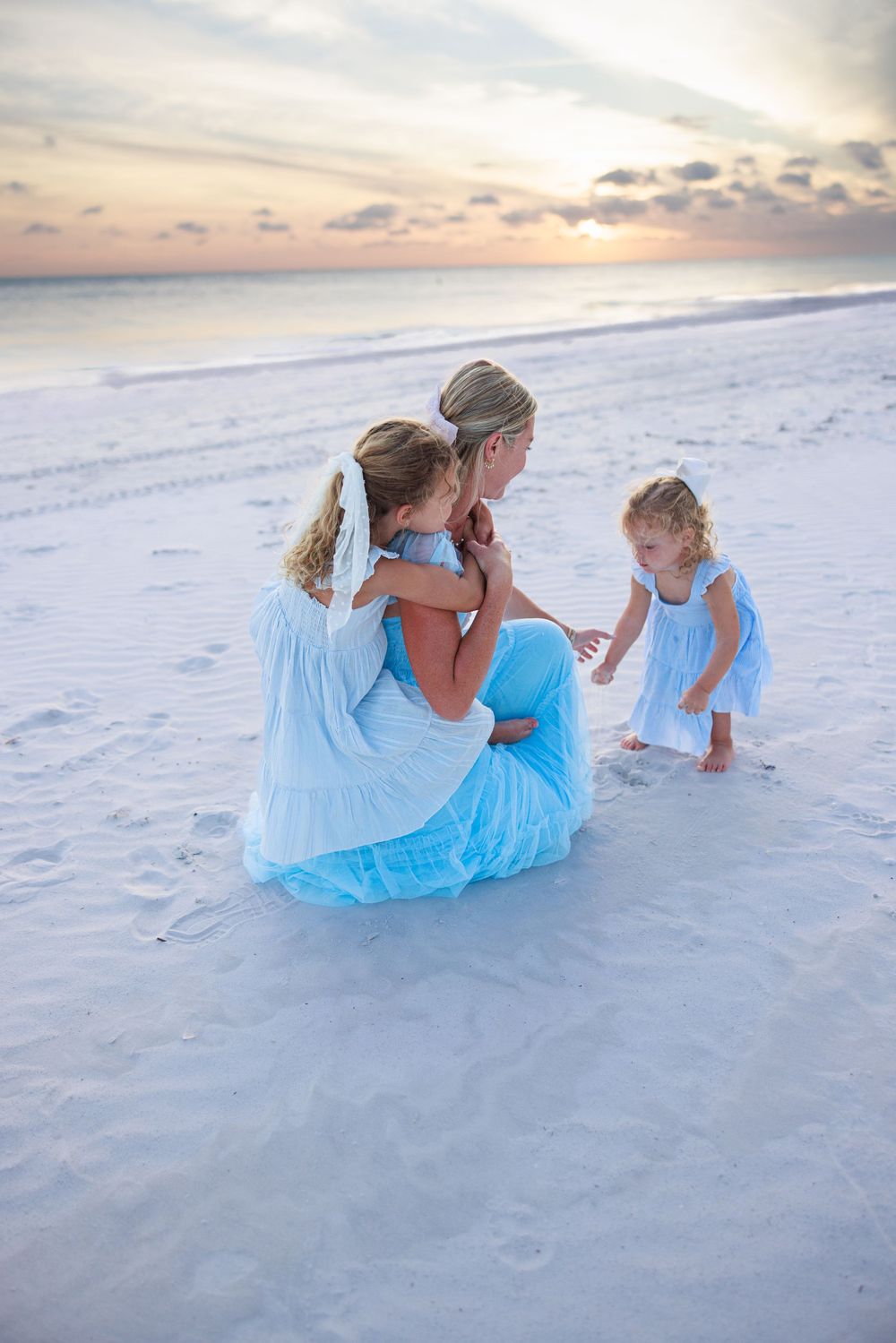 Family wearing light blue outfits plays together on a pristine white sand beach at sunset.