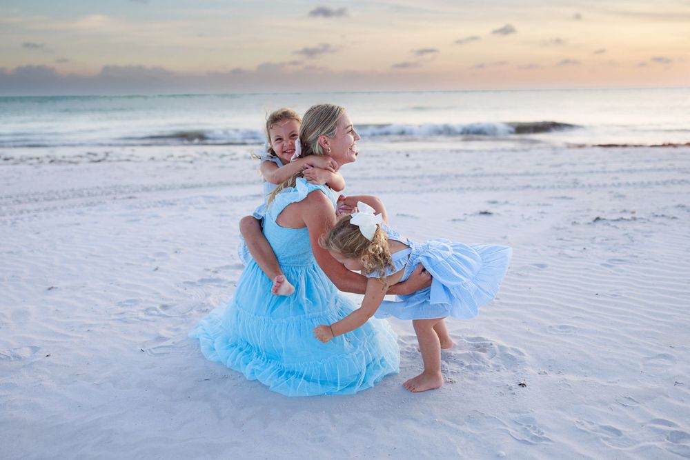 Family members wearing matching light blue dresses spin and play together on a white sandy beach at sunset.