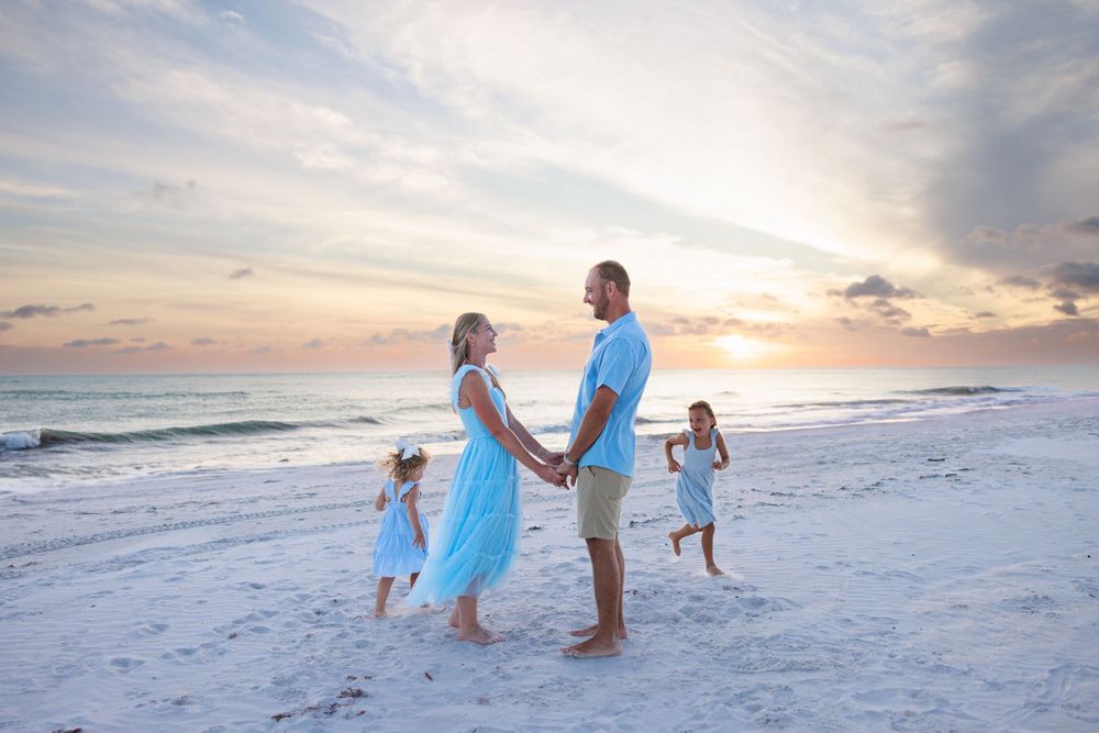 A family in light blue outfits dances together on a white sandy beach at sunset.