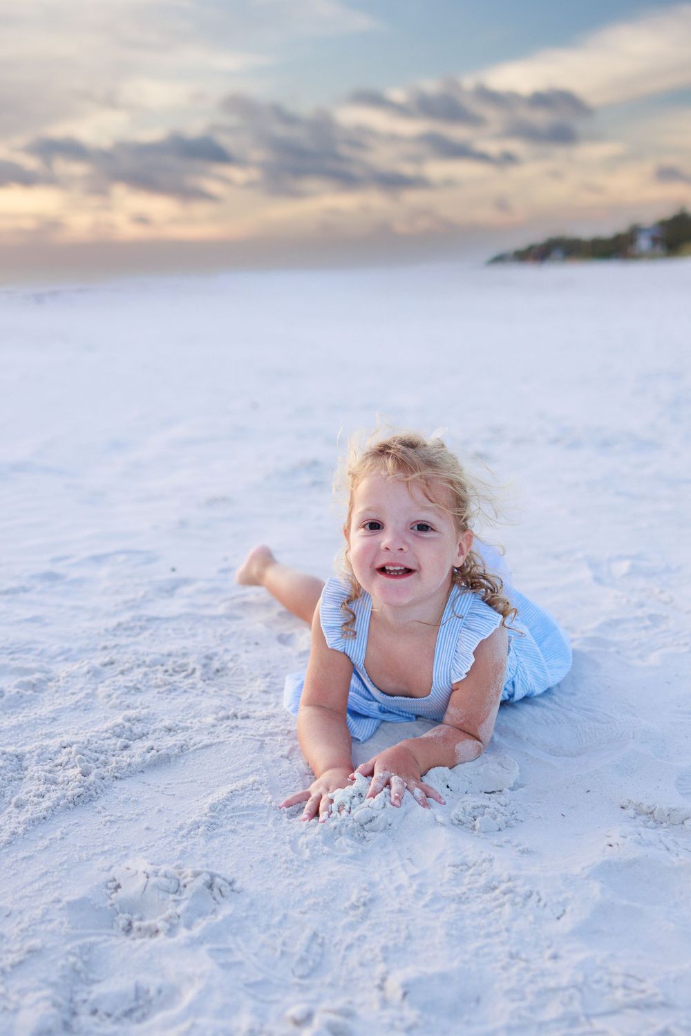 Young child wearing a light blue dress crawls and plays on pristine white sand beach at sunset.