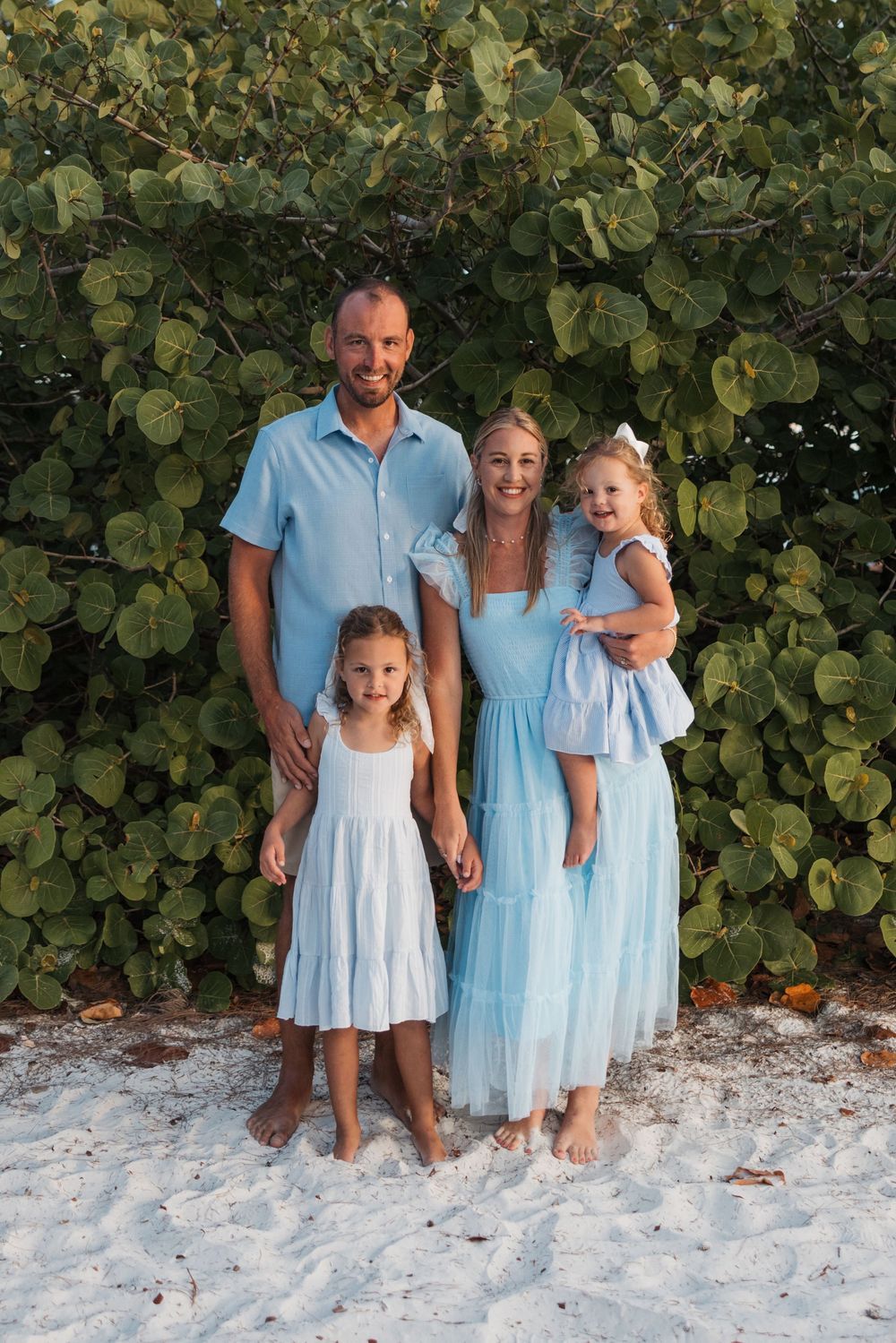 Group photo on a sandy beach with lush green foliage backdrop, wearing coordinated light blue and white outfits.
