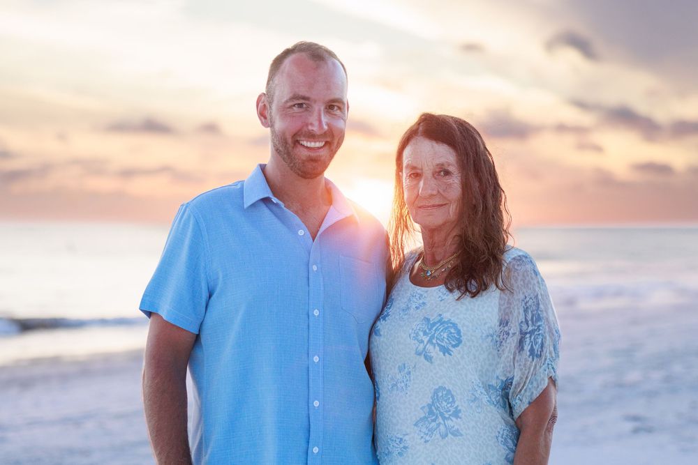 A couple stands together on the beach at sunset, with the man wearing a blue shirt and woman in a white patterned top.