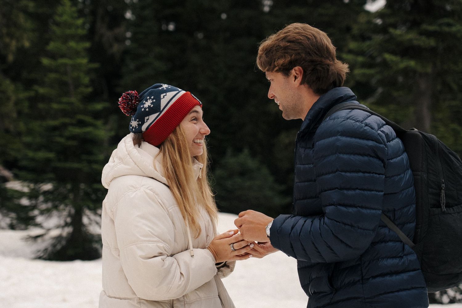 Two people share an intimate moment while holding hands in the snow, surrounded by evergreen trees in winter.