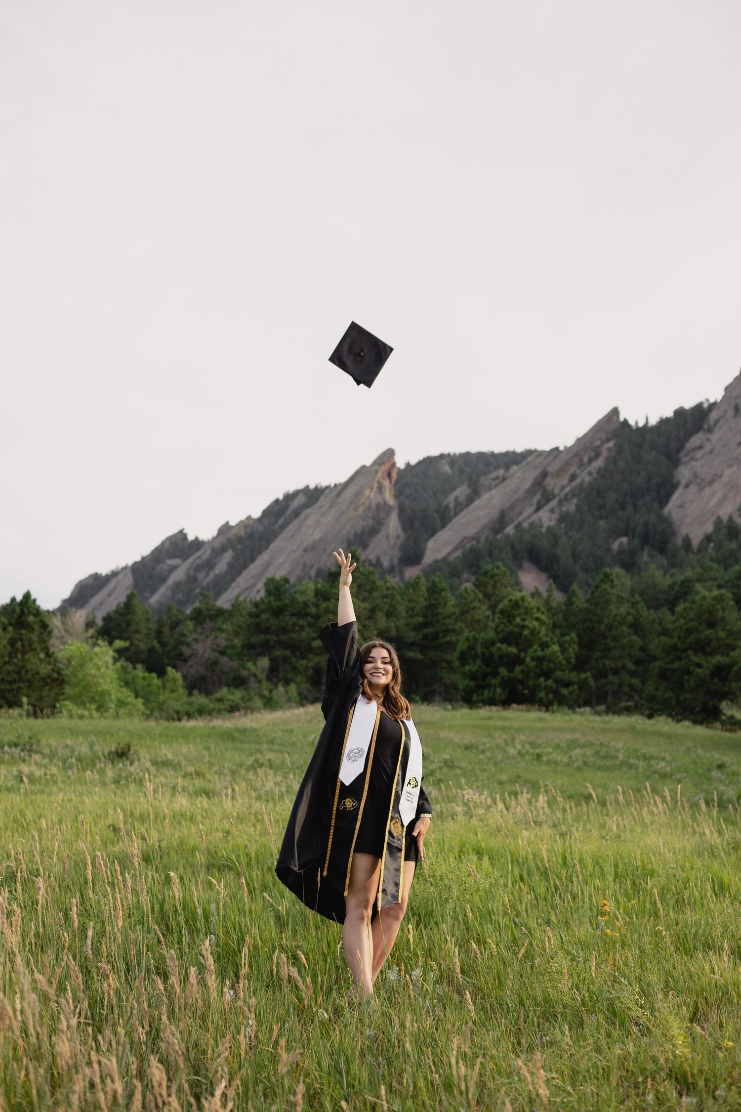 Graduate in cap and gown throws graduation cap in front of scenic mountain landscape with dramatic rock formations.