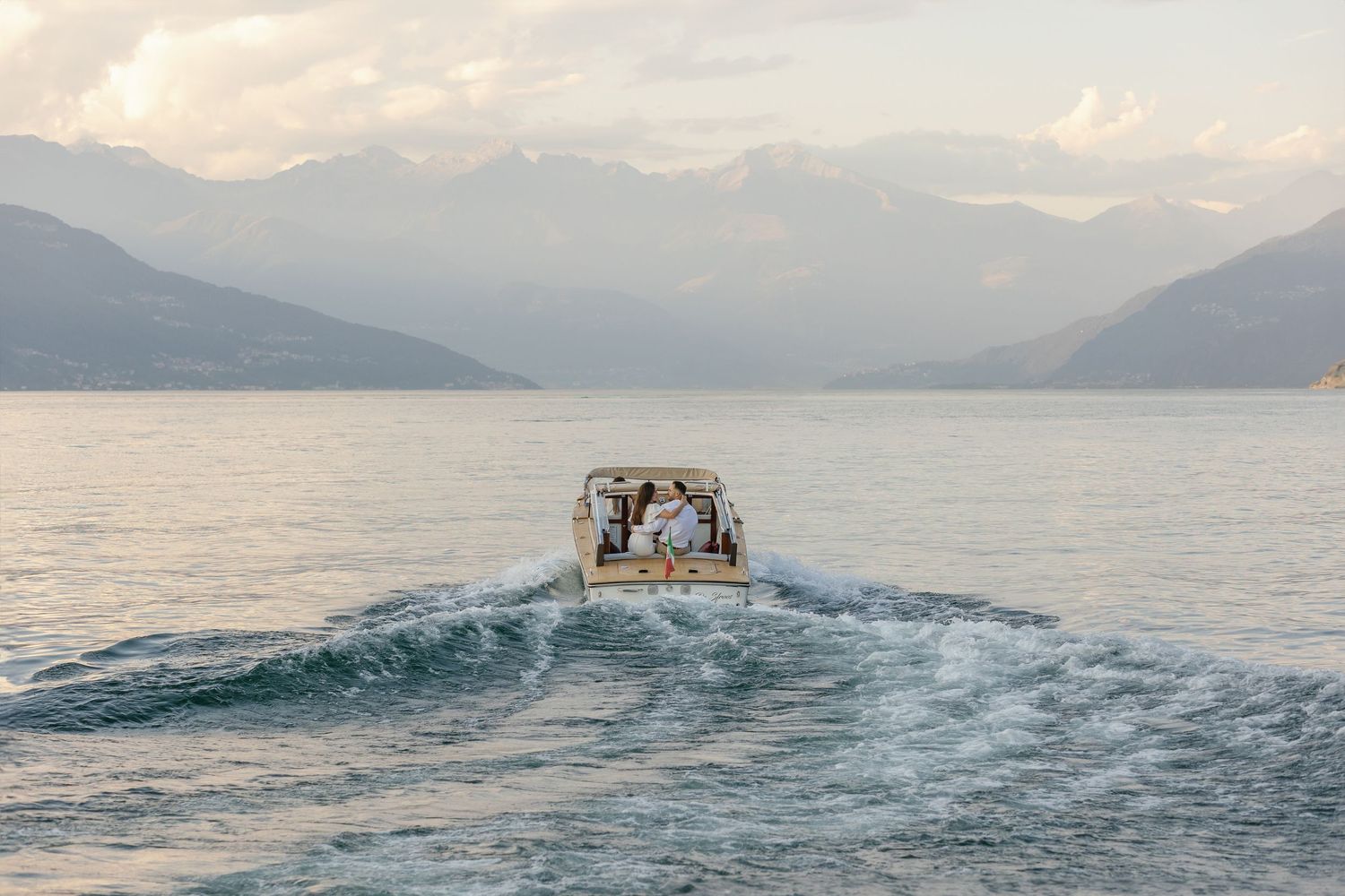 A wedding couple on a boat, creating a wake, with serene mountains and lake Como in the background.