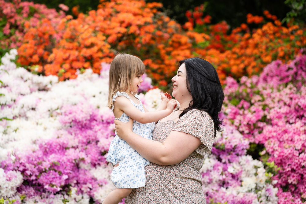 Mom and daughter smiling at each other in front of the rhododendron flowers.
