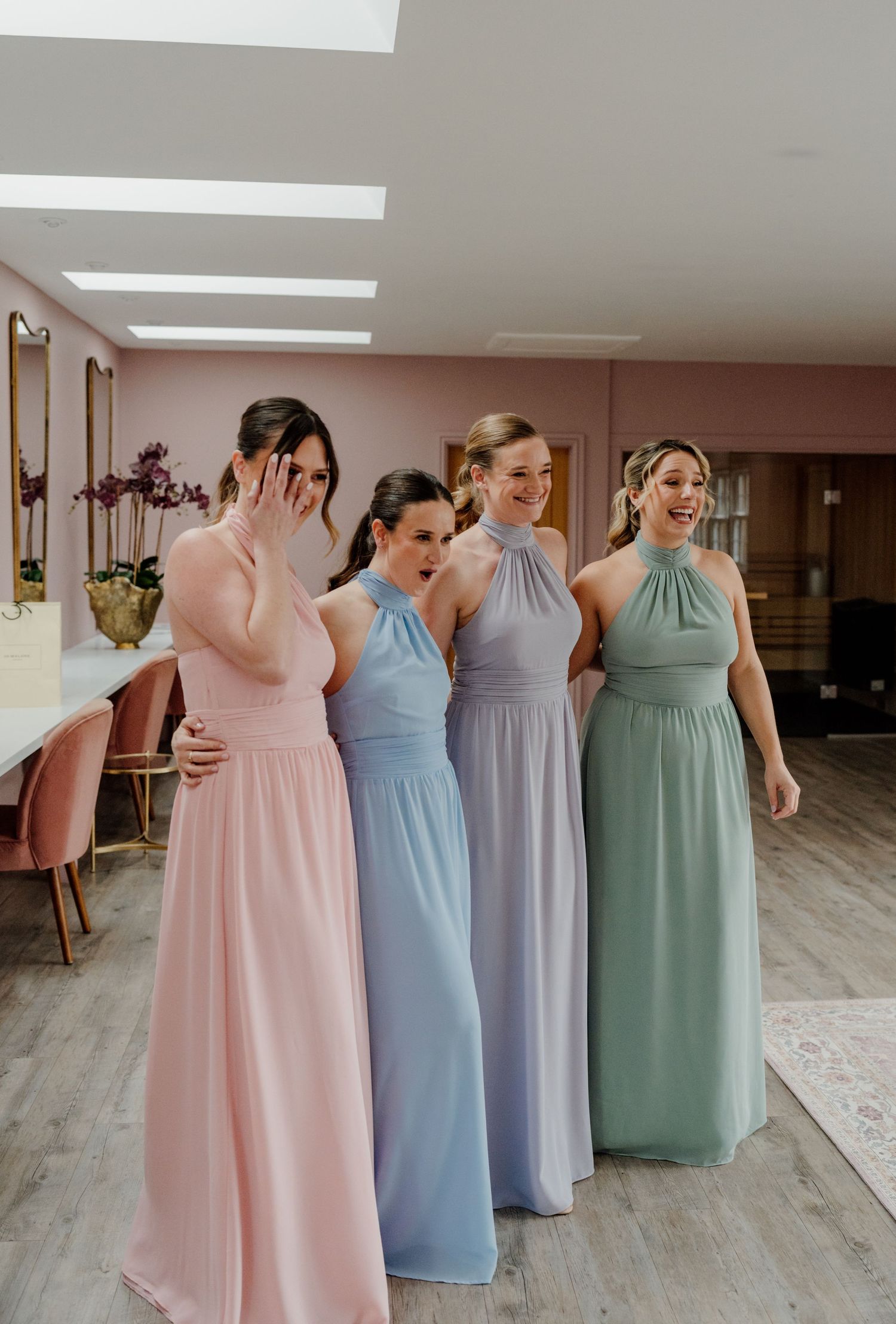 Bridesmaids in pastel pink, blue, lavender and sage green dresses laugh together at Quantock Lakes wedding Venue in Somerset
