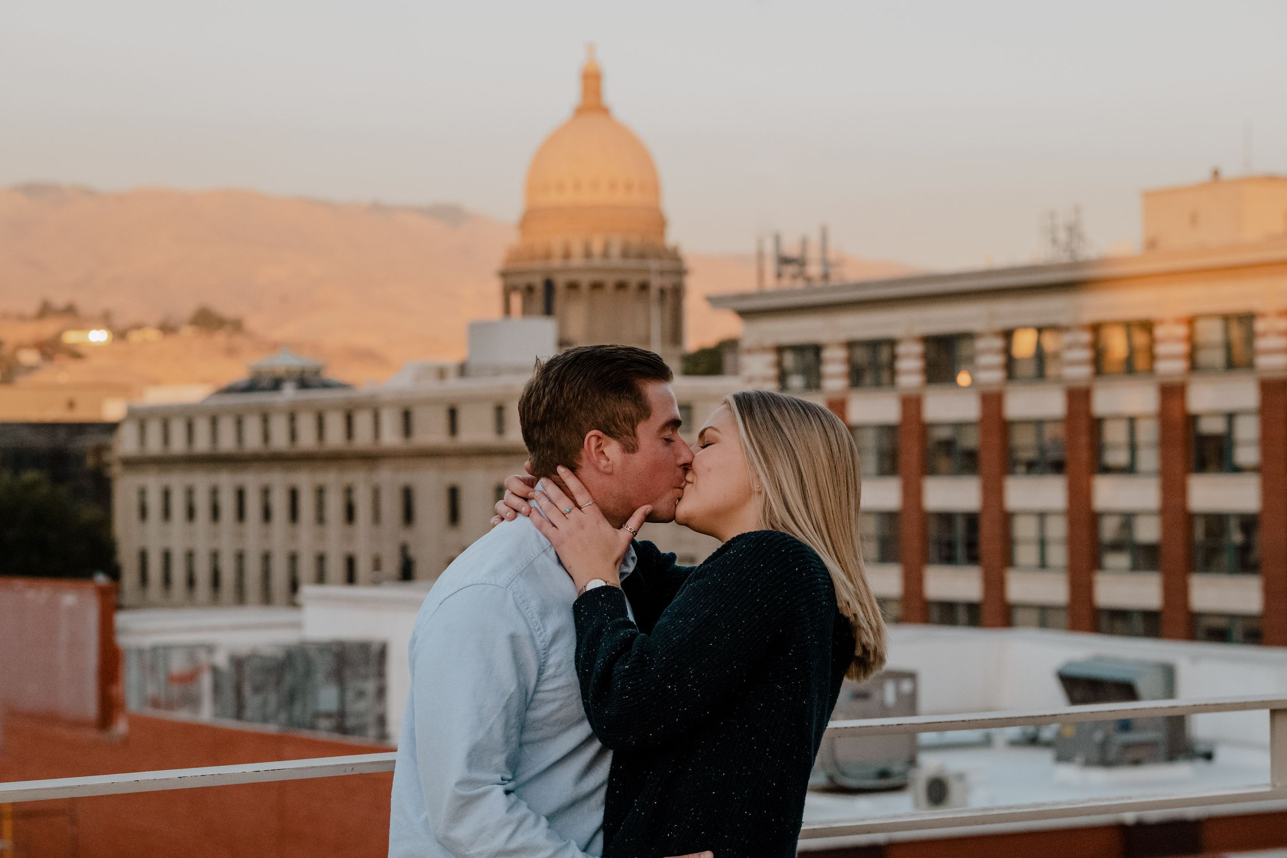 Romantic couple sharing a kiss on rooftop with Idaho State Capitol building dome visible in sunset background in Boise.