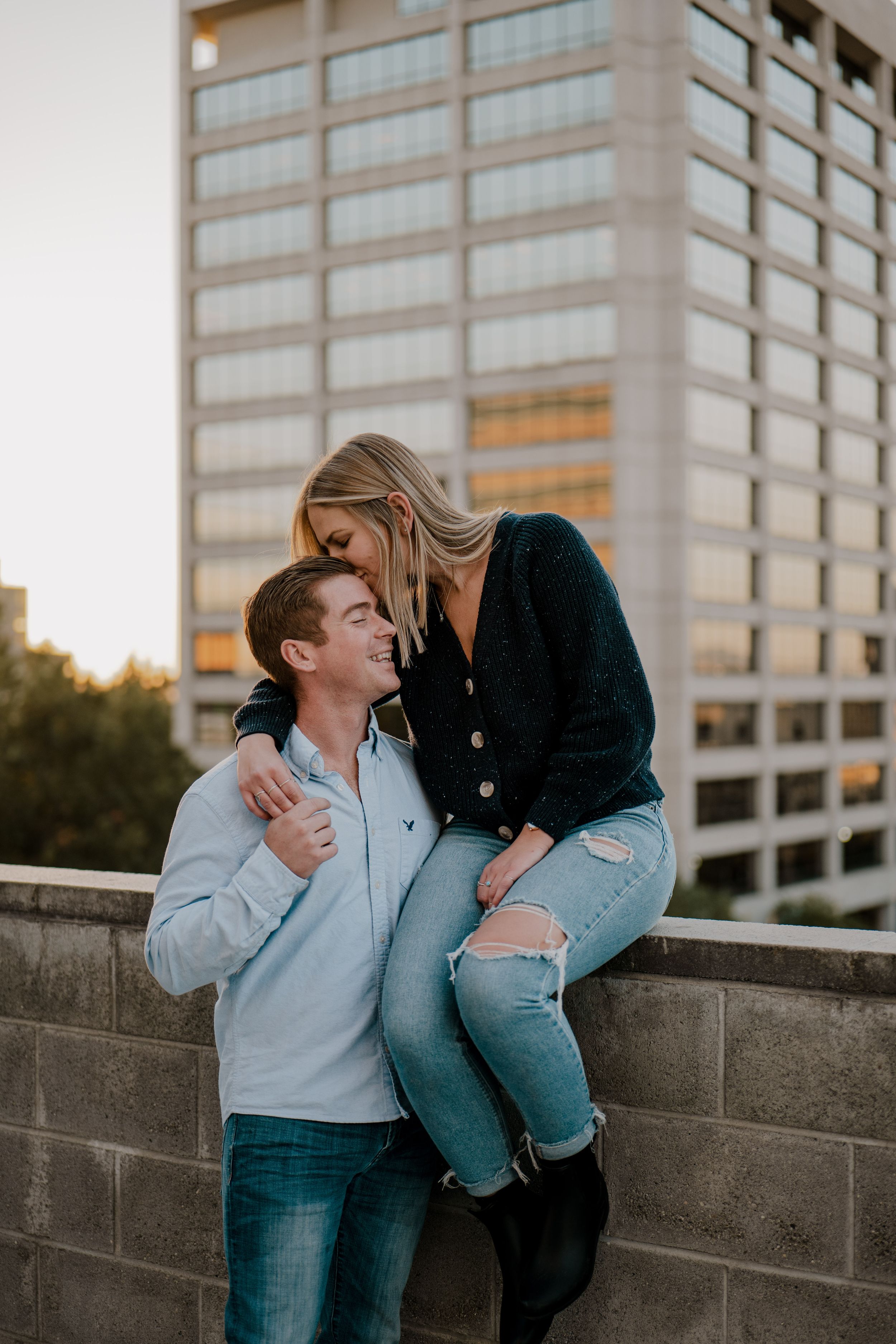 A romantic couple shares an intimate moment at sunset against an urban backdrop with a modern office building.
