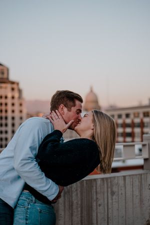 A romantic couple sharing a passionate kiss on a rooftop with city buildings and a domed capitol in the background at sunset.