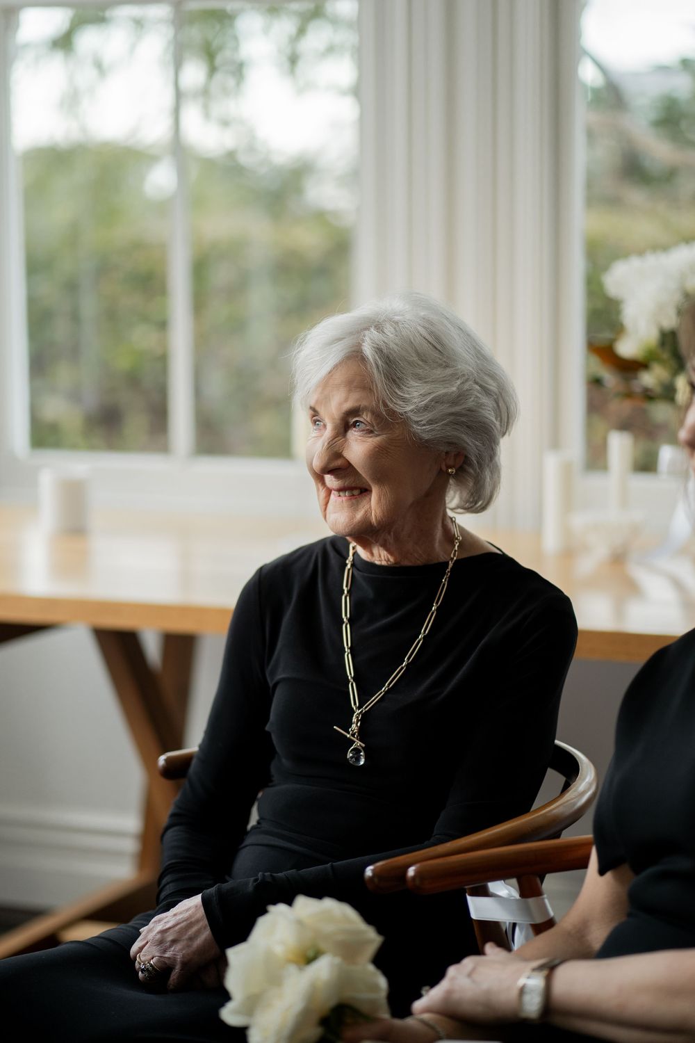 Elegant senior woman in black sitting by a sunny window at formal event.
