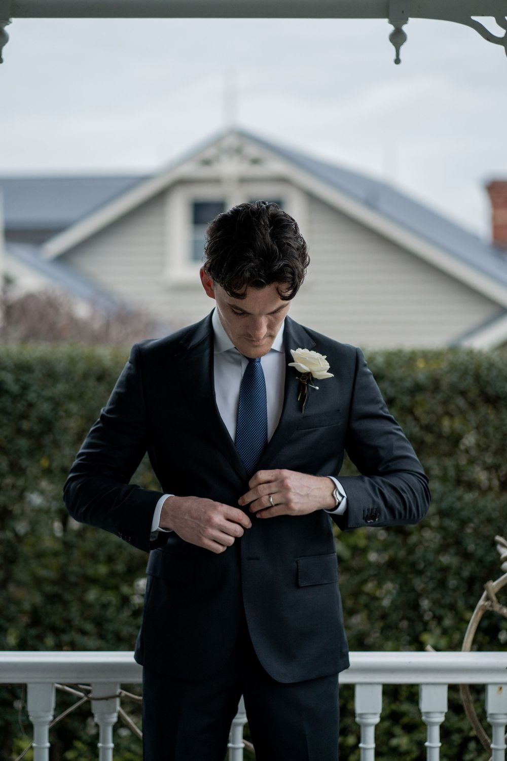Groom in dark suit adjusts clothing while standing on porch with white railings.
