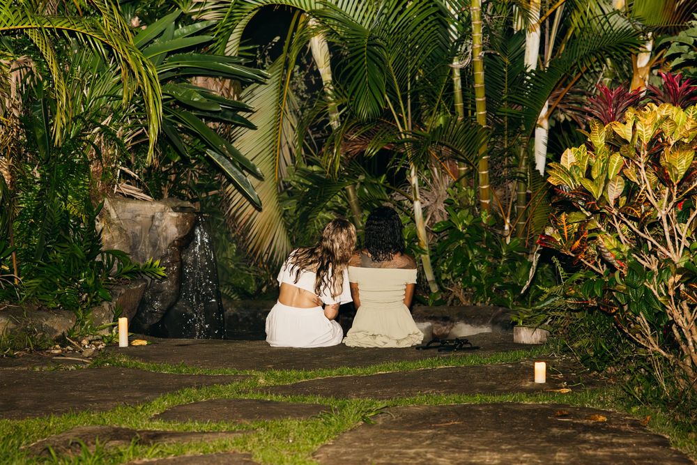 Two people sitting on pathway surrounded by tropical garden at dusk.