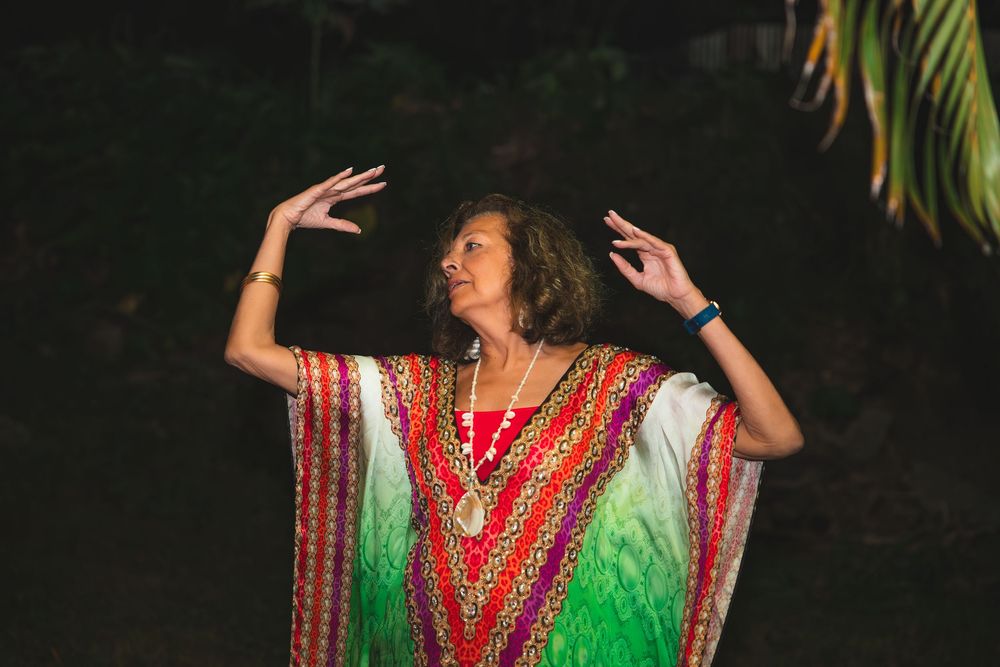 Person wearing a colorful boho-style dress with ornate patterns dances outdoors at night with palm trees in background.