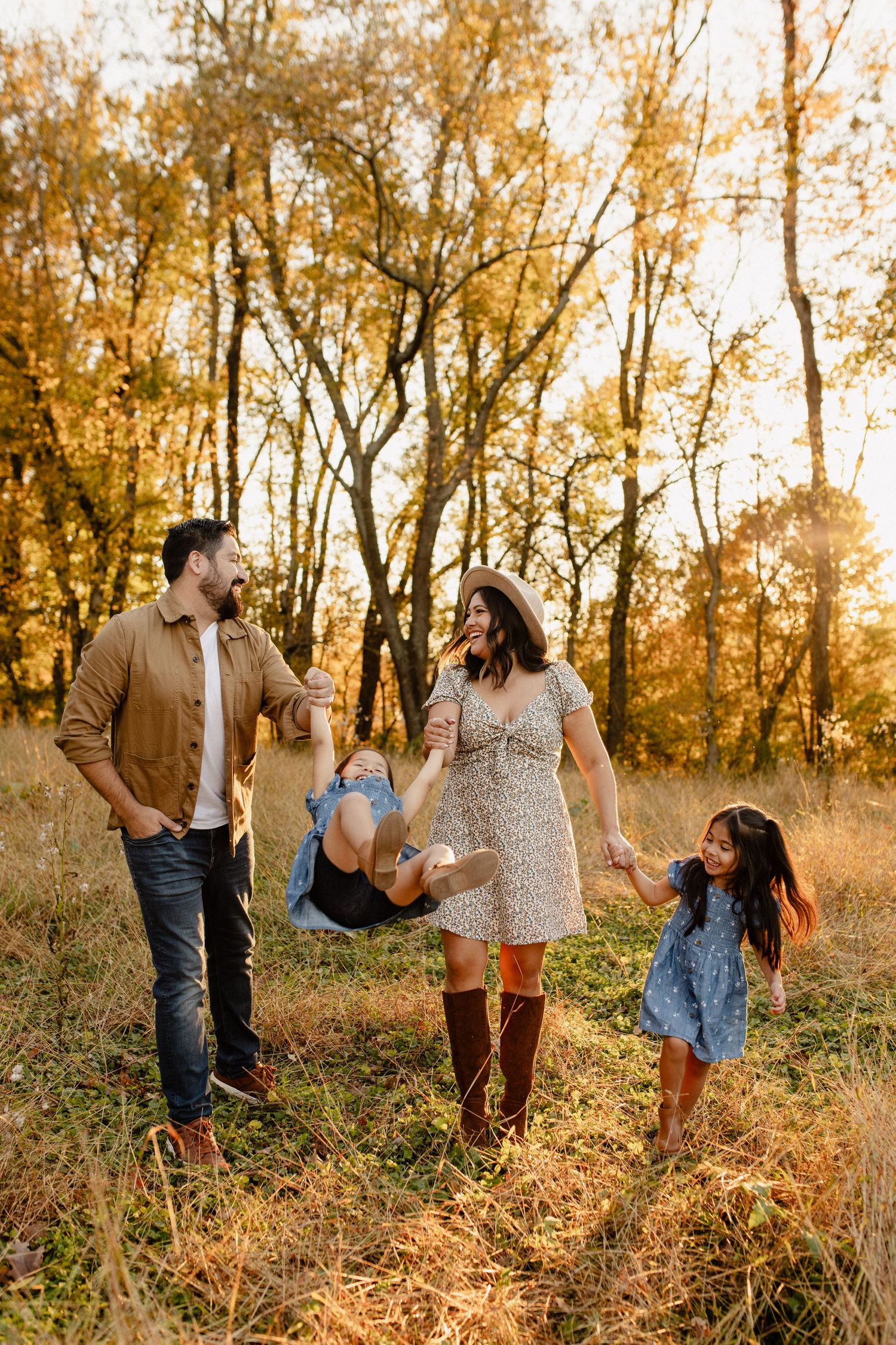 Happy family enjoying a walk together in a golden autumn forest at sunset while swinging their young child shot by Maryland Photographer
