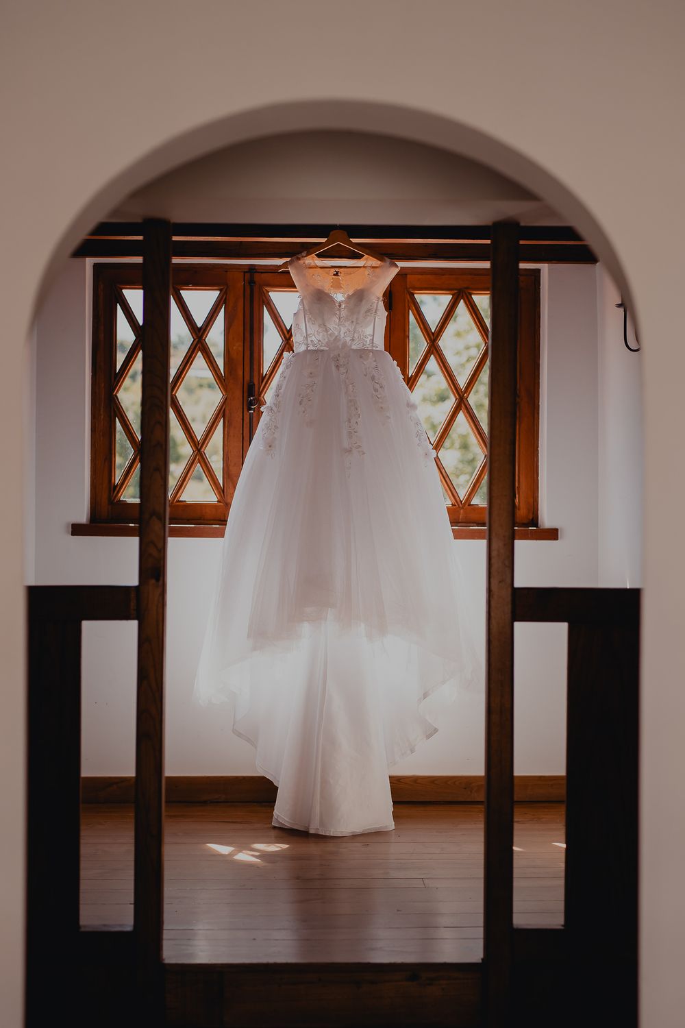 Elegant white wedding dress hanging in an arched doorway with diamond-paned windows.
