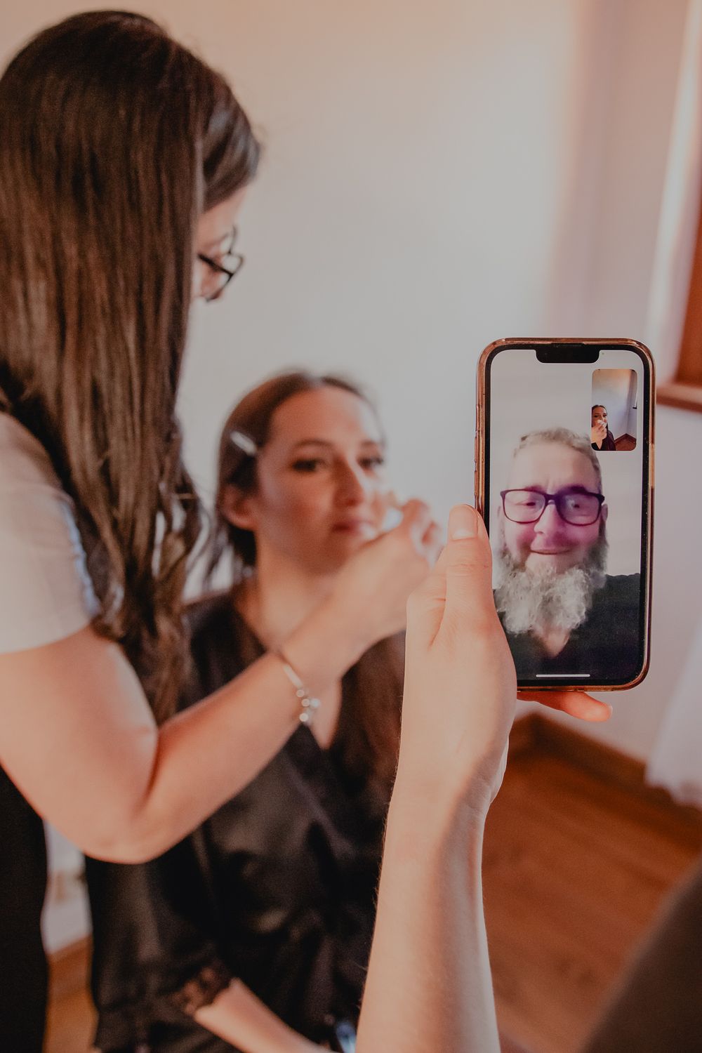 A makeup artist works on a client while someone holds up a phone displaying a video call.